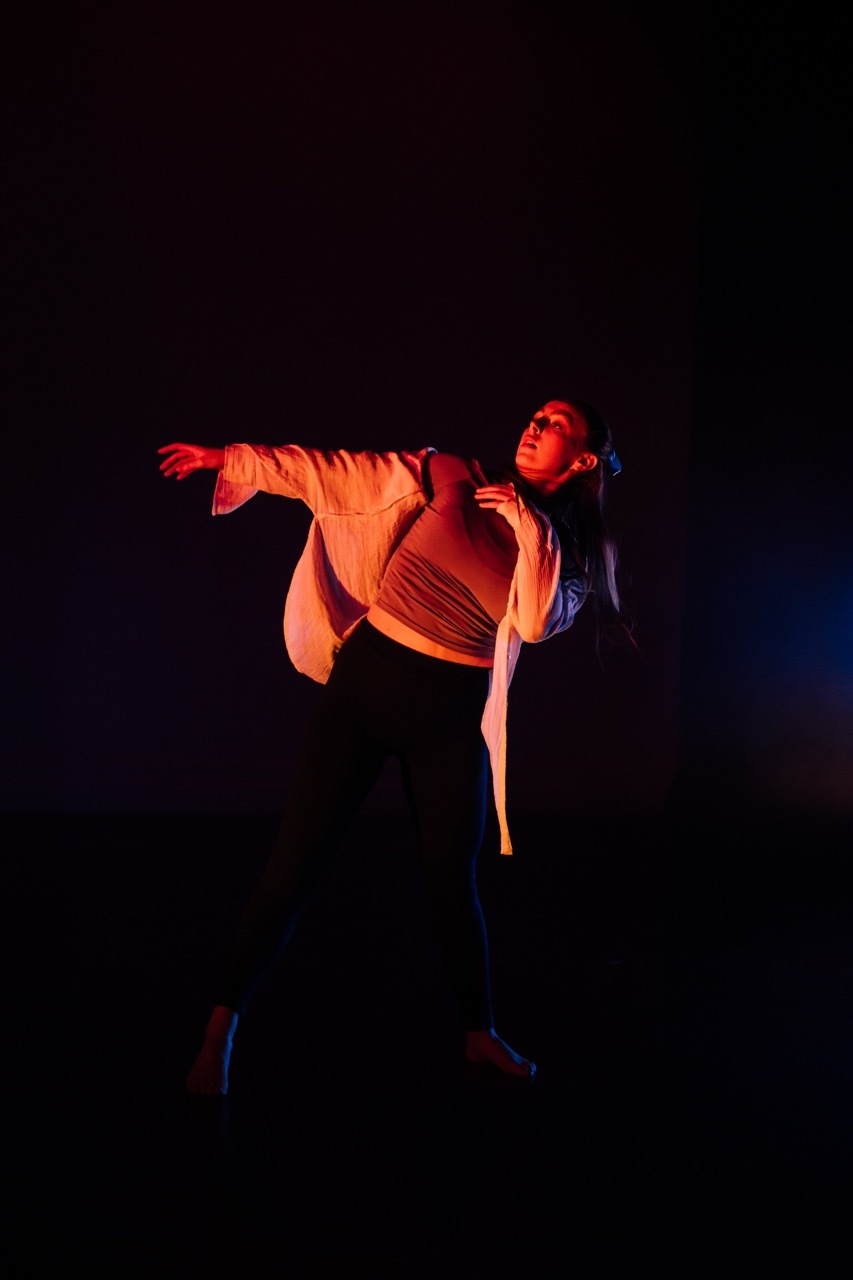 Dancer in mid-leap against dark background, illuminated by warm orange and red stage lighting, arms extended outward.