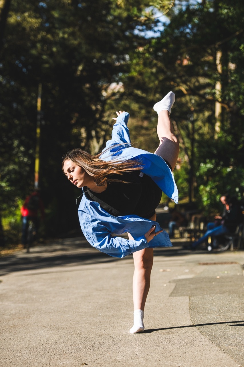 Woman in blue denim jacket and white trainers performing breakdance move on one leg in park, hair flowing, trees in background.