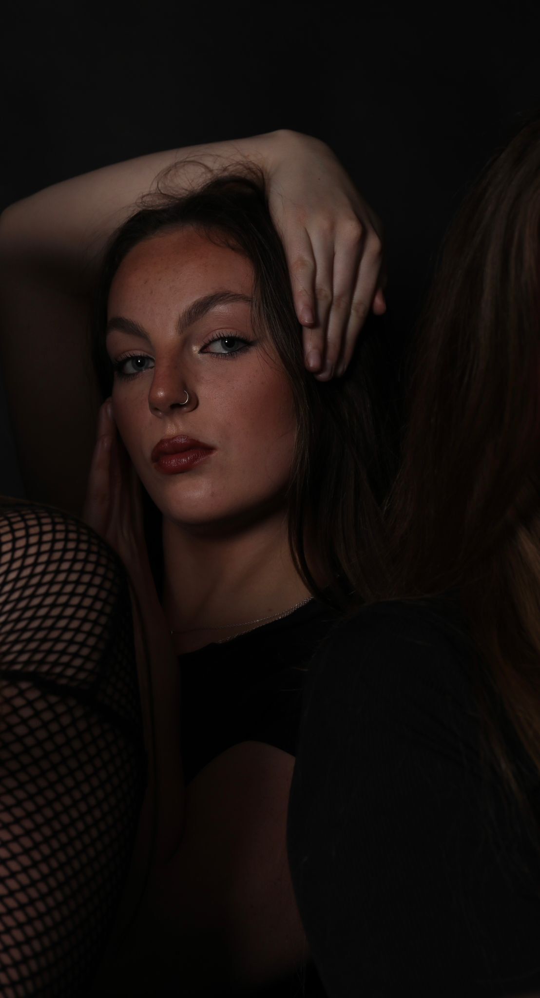 Woman with blonde hair and dark makeup holds hand to forehead, wearing black top with white fishnet overlay in dim lighting.