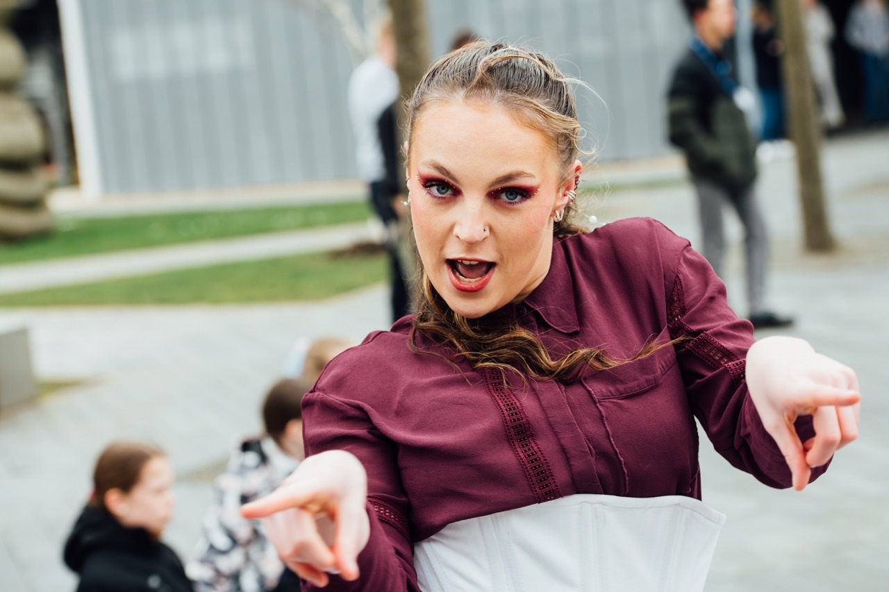 Woman in burgundy top with arms outstretched, mouth open, leaning forward towards camera in public square setting.