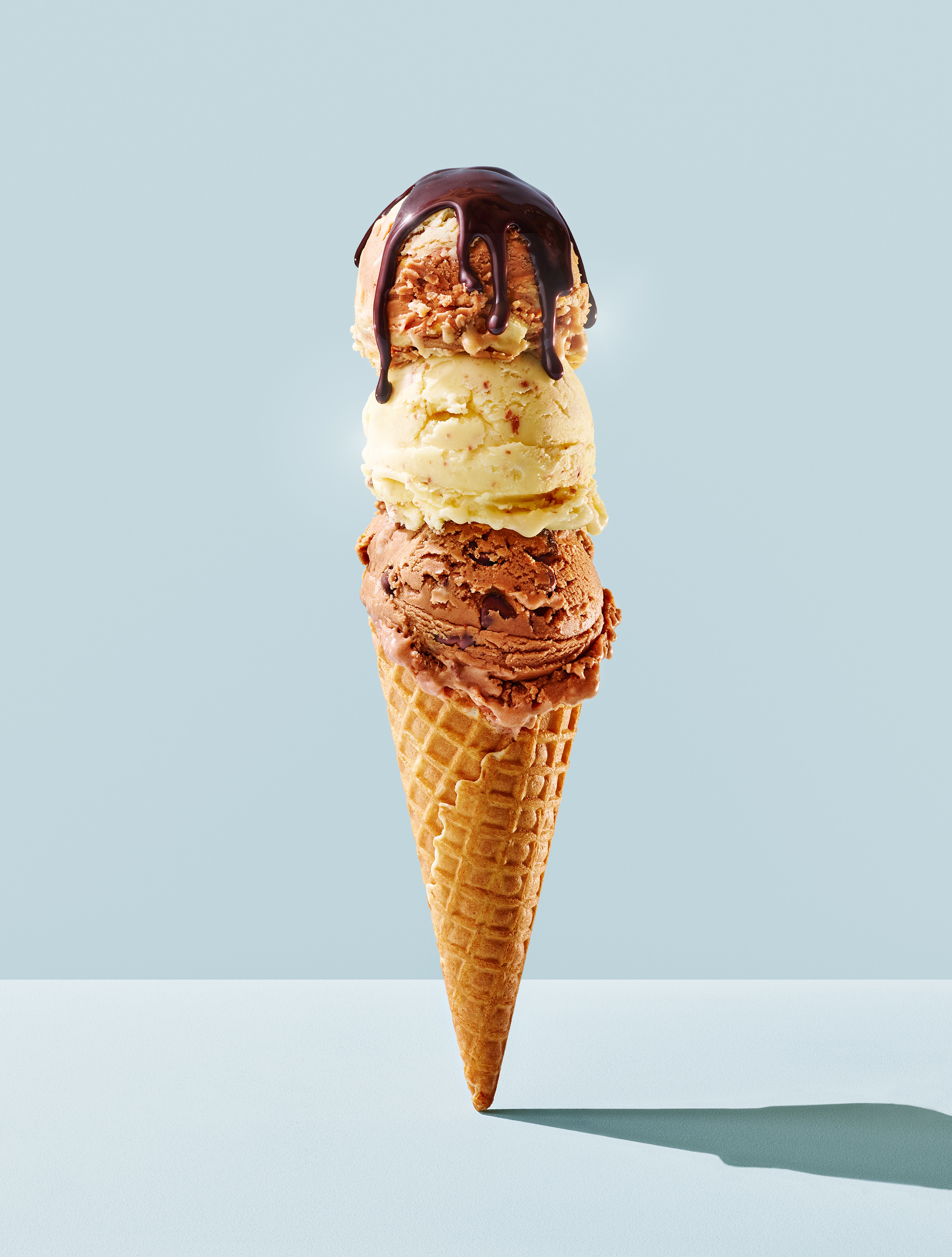 An ice cream cone with three scoops stacked on top of one another, topped with chocolate sauce.