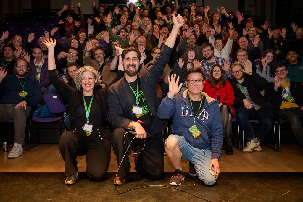 A group of people seated in an auditorium raise their hands, with four people kneeling in the front row and lanyards visible.