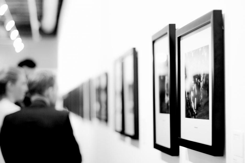 Close-up of a gallery wall with a row of framed artworks going down. People can be seen looking at some of these artworks in the background.