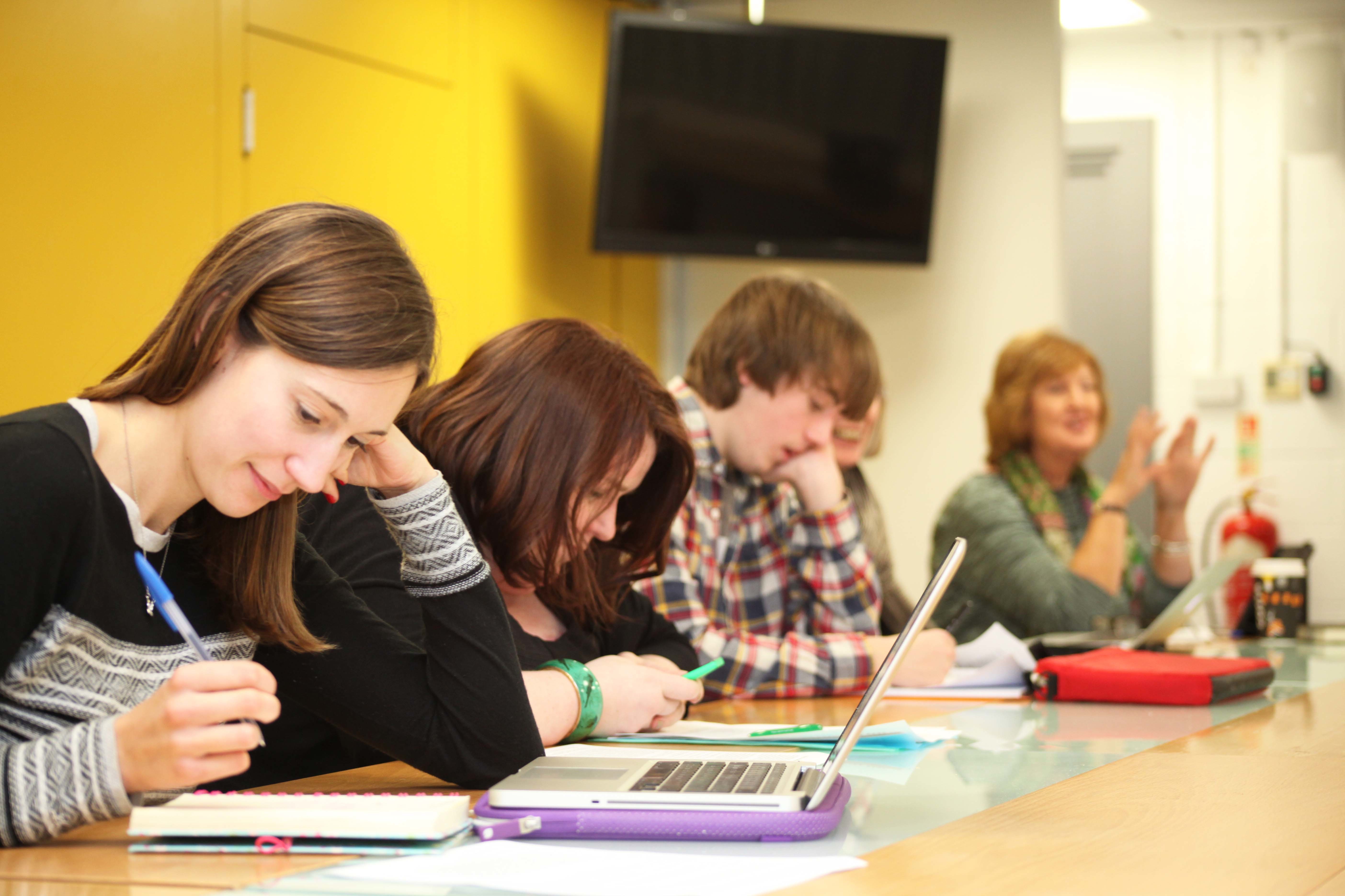 A line of students sitting at tables taking notes in notebooks, on phones and on laptops.