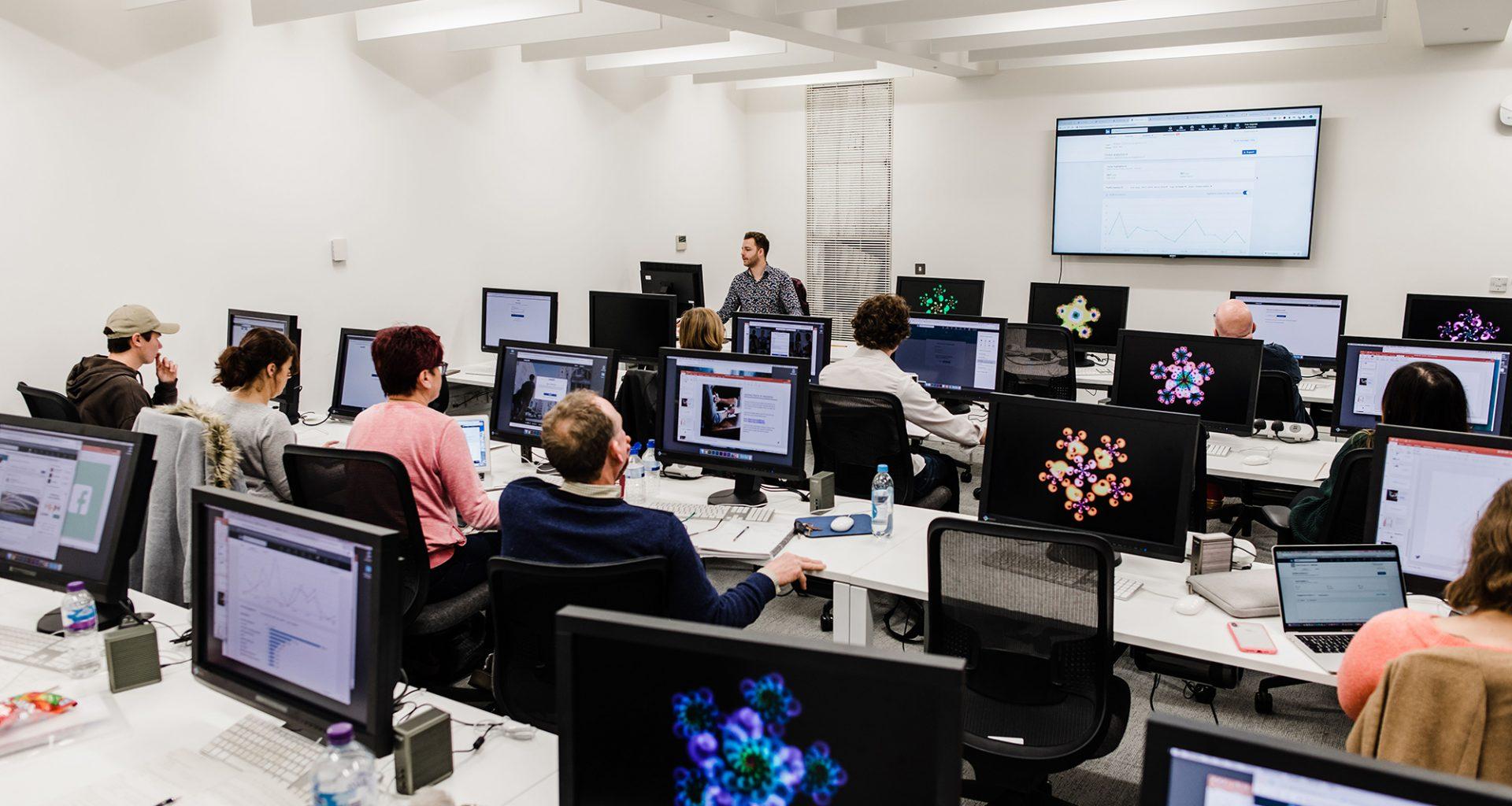 Students sitting at banks of desks with computers on them. A lecturer gives a presentation at the front.