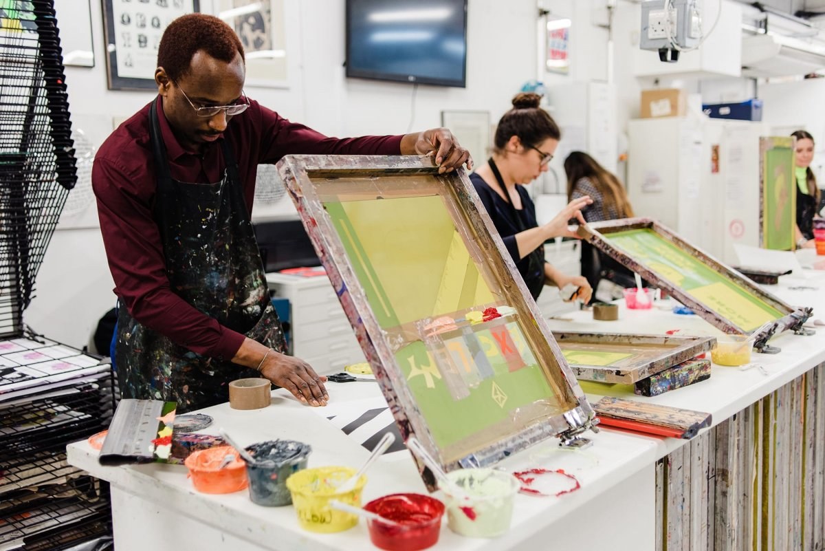 Two students working at a long table in a studio holding screen printing equipment.