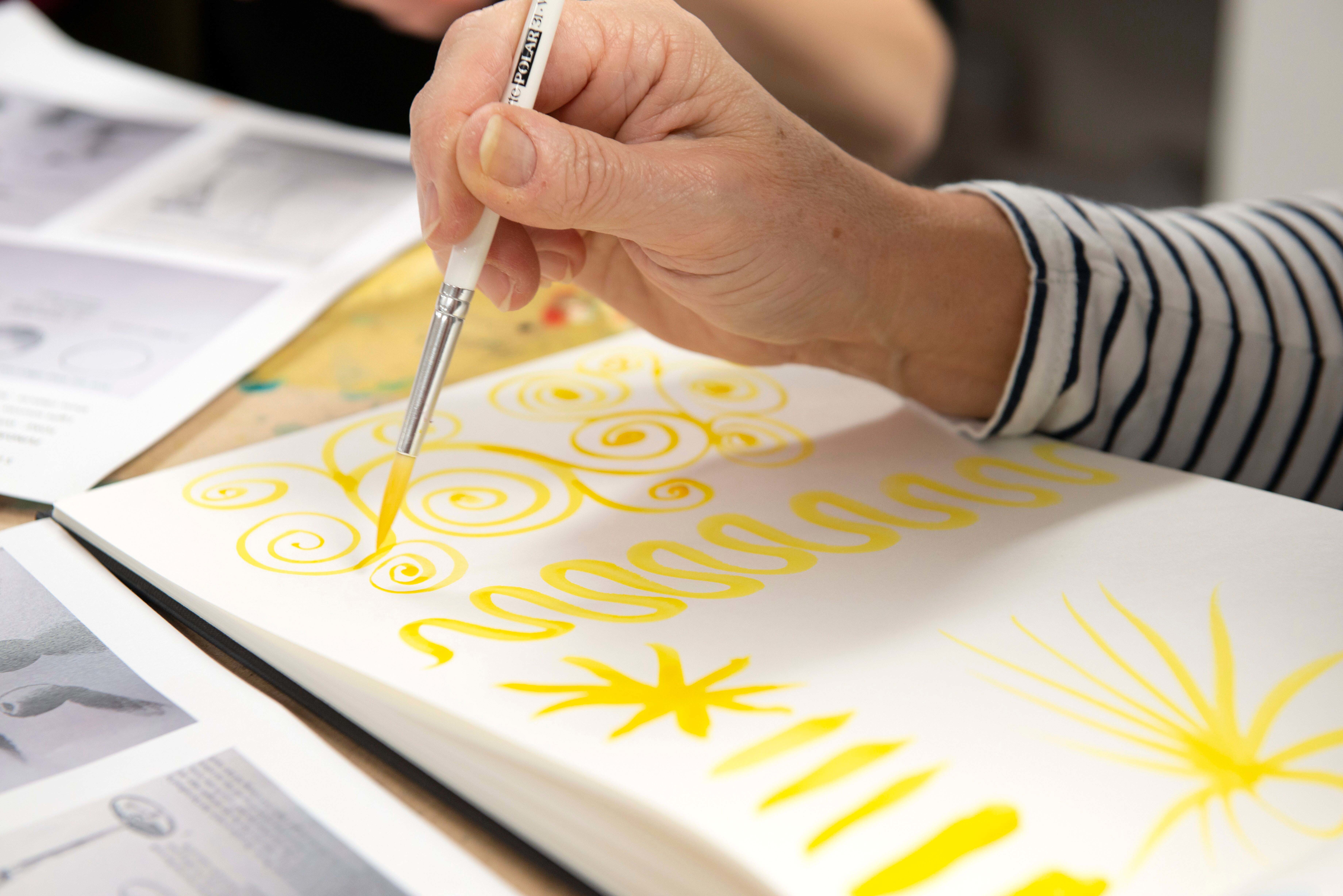 Student paints a curlicued pattern on a sketchbook page in yellow paint. Other lines and star patterns have been painted next to it.