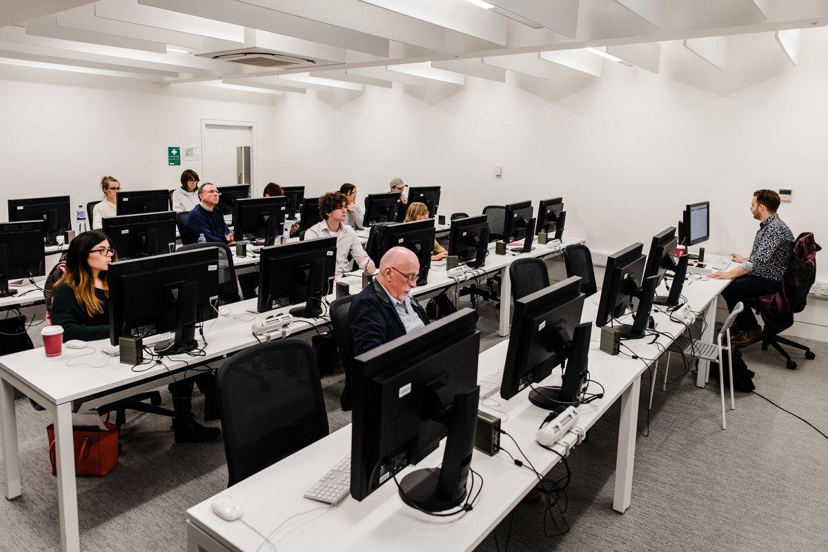Banks of desks with computers on them and several people working.