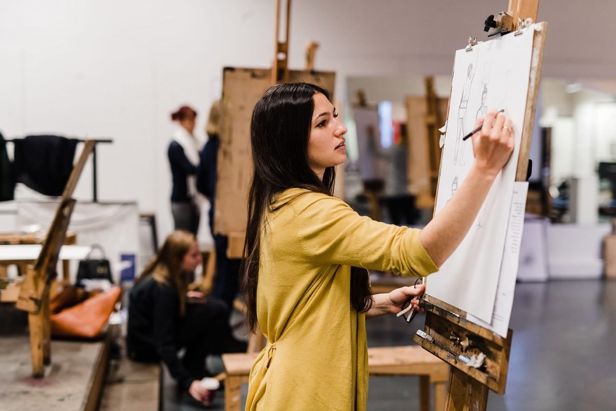 A student standing at an easel and drawing in pencil as part of a life drawing session.
