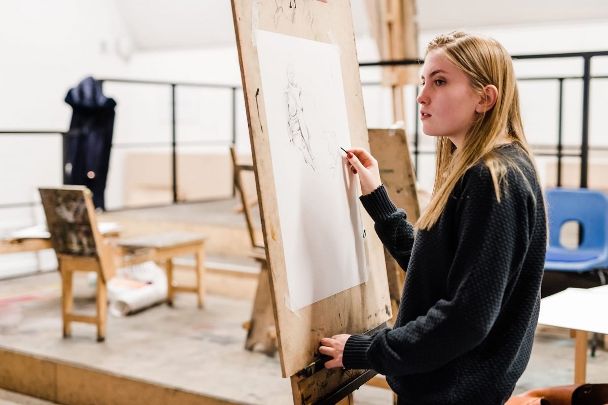 A student standing at an easel and studying something off camera, drawing a human figure in pencil as part of a life drawing session.