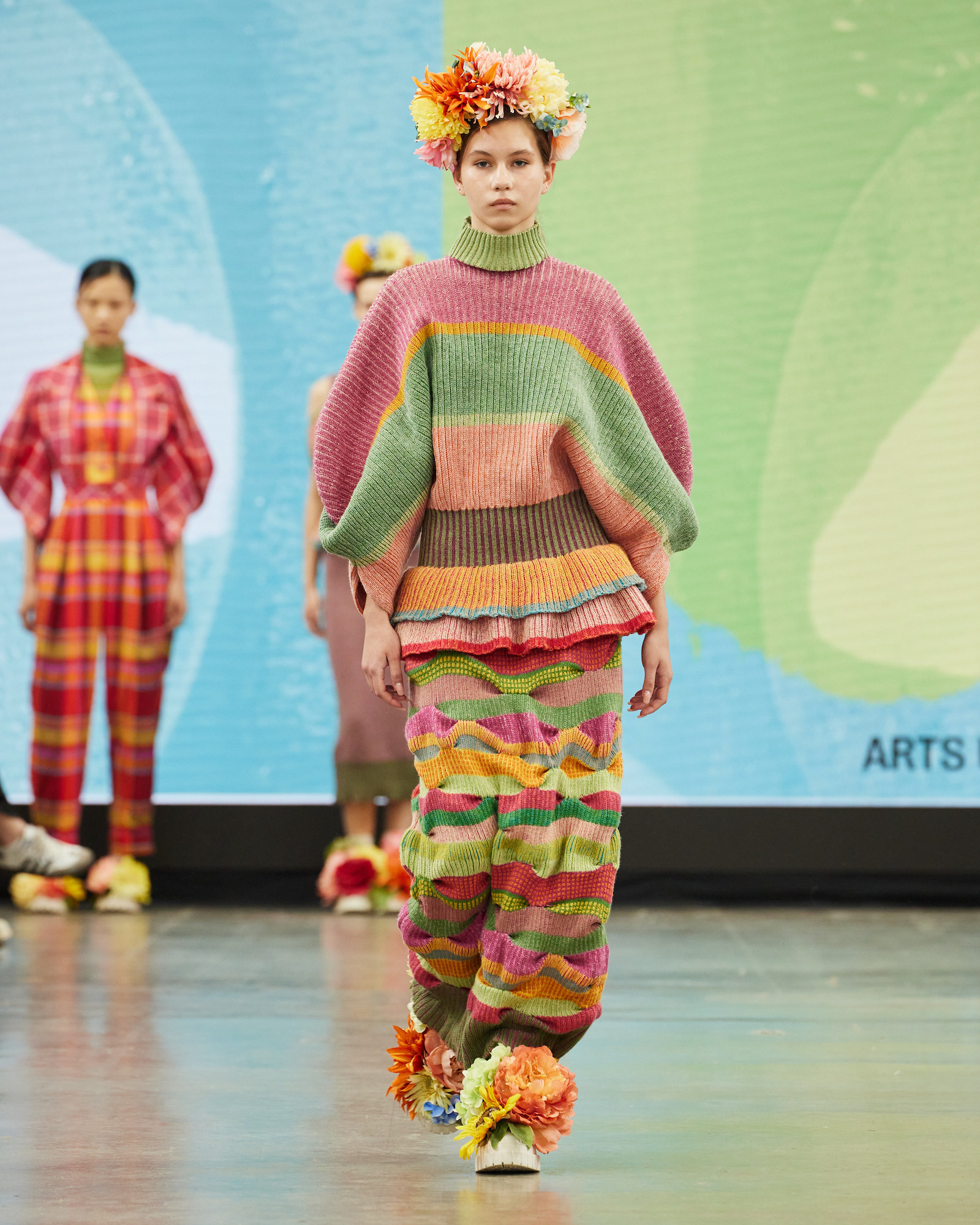 A model walking down the catwalk at Graduate Fashion Week 2024 wearing a colourful design by a BA (Hons) Fashion student. Shoes and headdress have flower accessories.