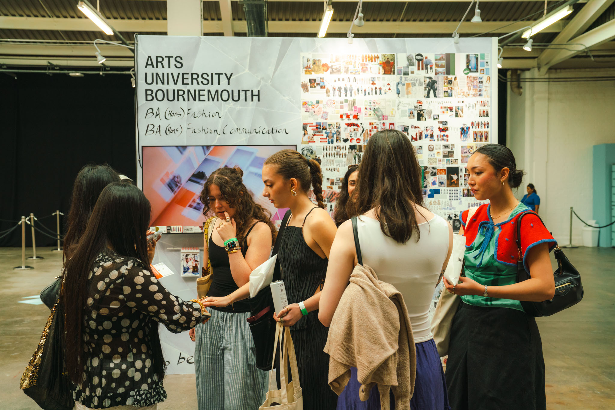 Group of students standing near Arts University Bournemouth exhibition display with white banner and collage of images in industrial venue.