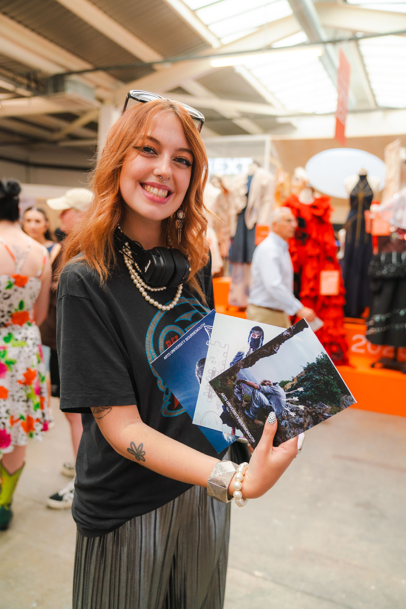 Woman with red hair holding small posters at GFW, wearing black top and pearl necklace, sunglasses on head.