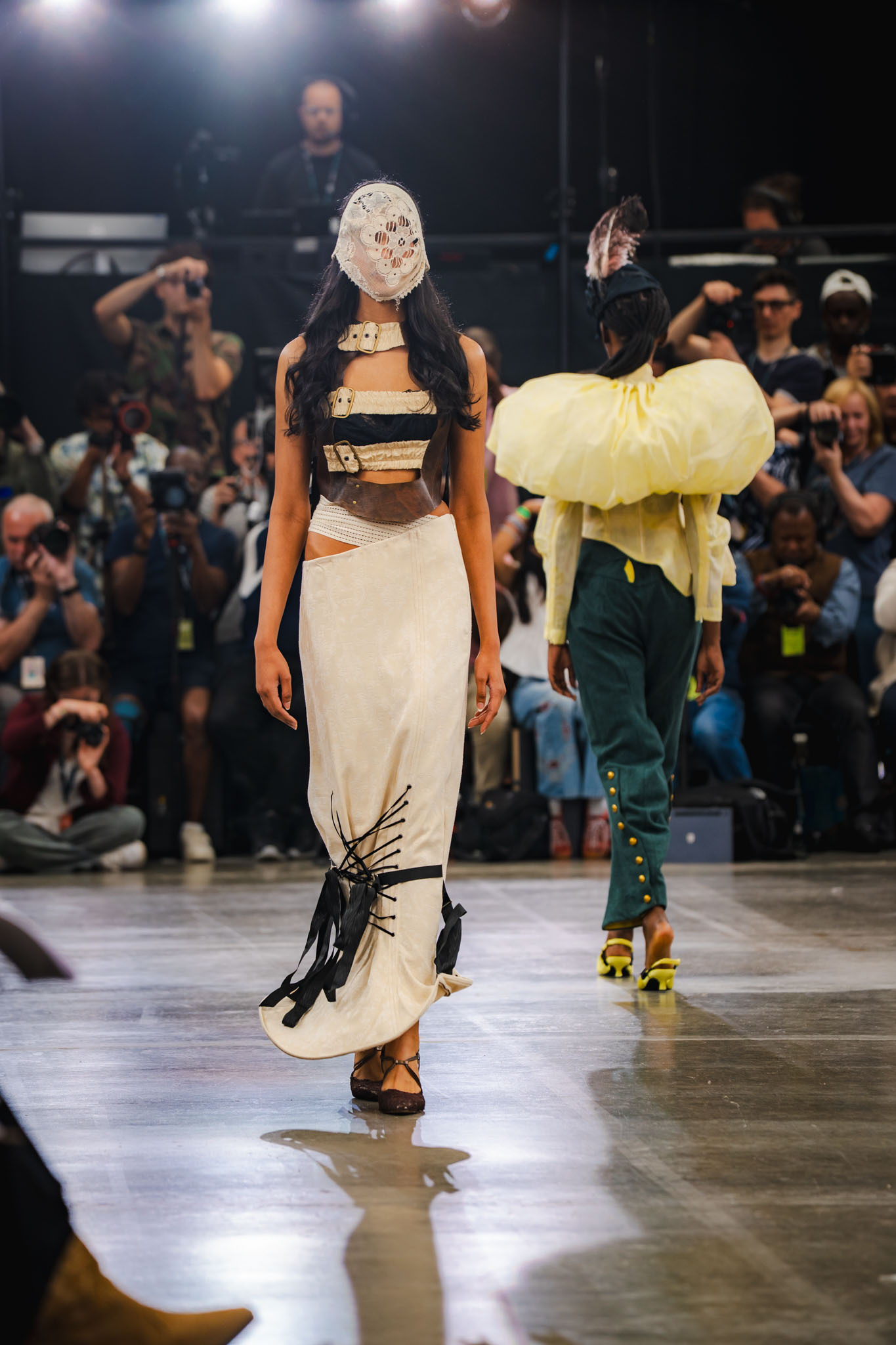 Two models on catwalk wearing avant-garde outfits: one in white dress with face covering, another in yellow ruffled headpiece and green trousers.