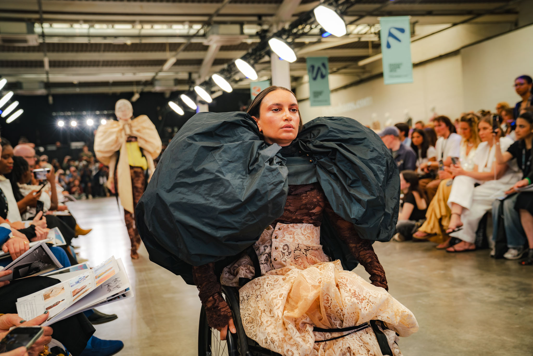 Model on catwalk wearing voluminous dark green puffy sleeves with textured beige lower garment, surrounded by seated audience in industrial venue.
