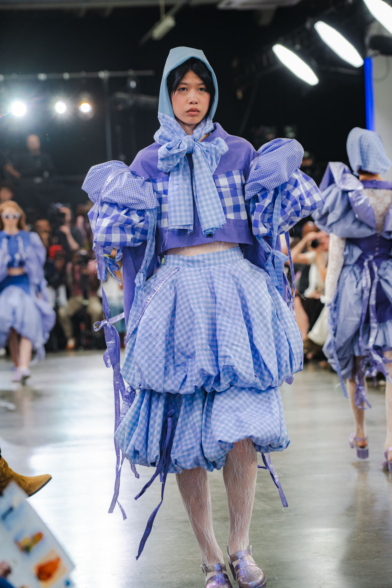 Model walks runway wearing layered blue and white gingham outfit with puffy sleeves, hooded top, and ruffled skirt elements.
