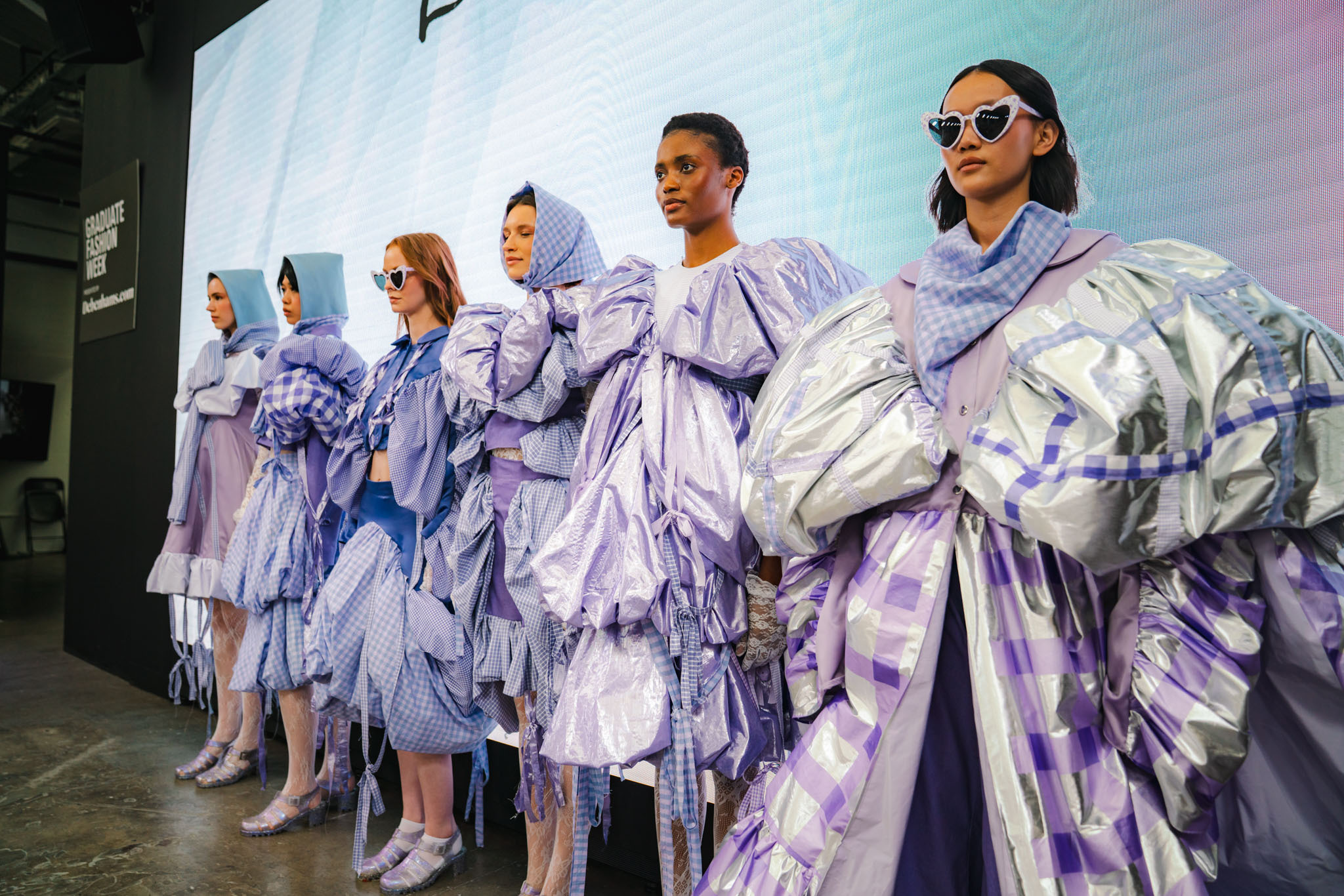 Five fashion models on runway wearing voluminous purple and white garments with dramatic sleeves and layered fabric details.