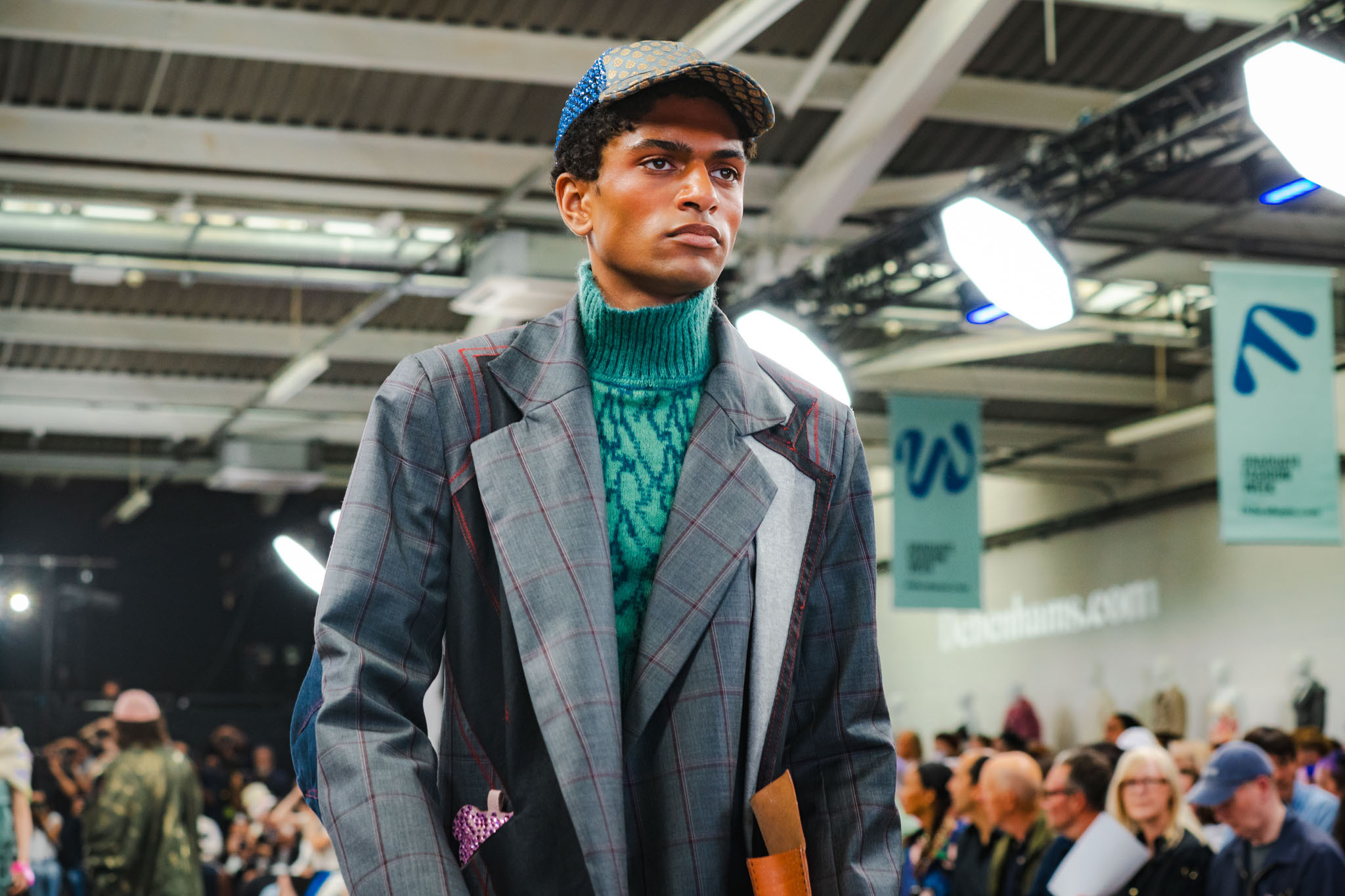 Male model on runway wearing grey oversized blazer and green turtleneck jumper, industrial ceiling with exposed beams above seated audience.