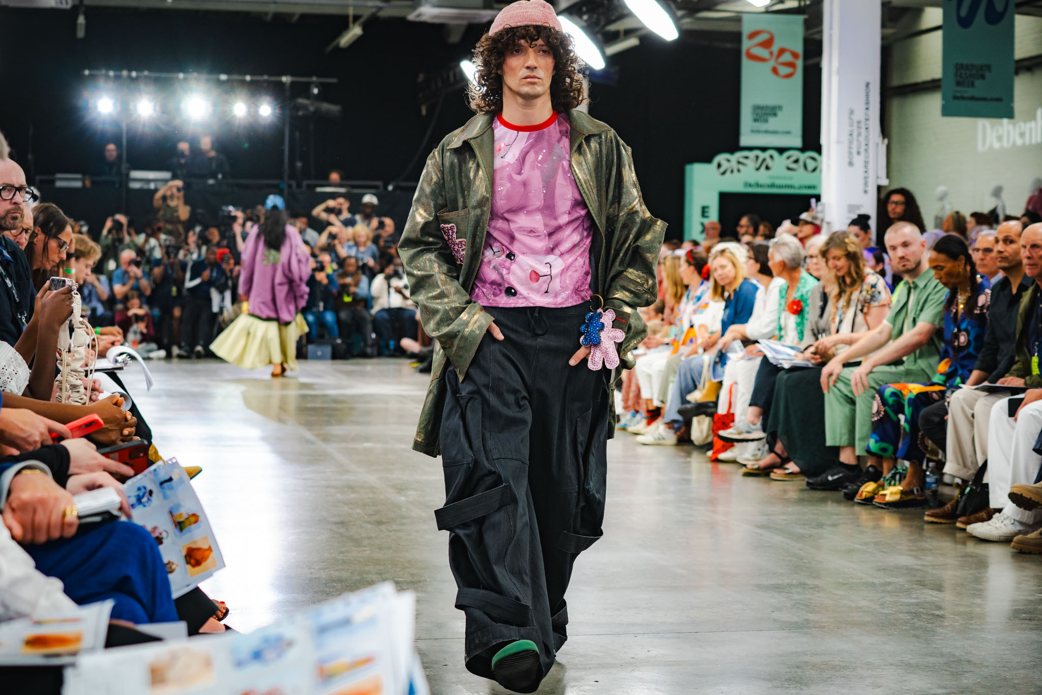 Model walks runway wearing pink cap, olive jacket, pink top, and black trousers whilst audience sits on both sides of catwalk.