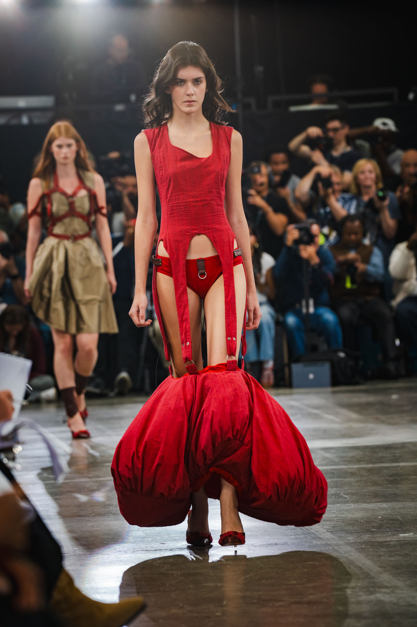 Model walks runway in avant-garde red gown with cut-out panels and voluminous bottom section, photographers and audience in background.