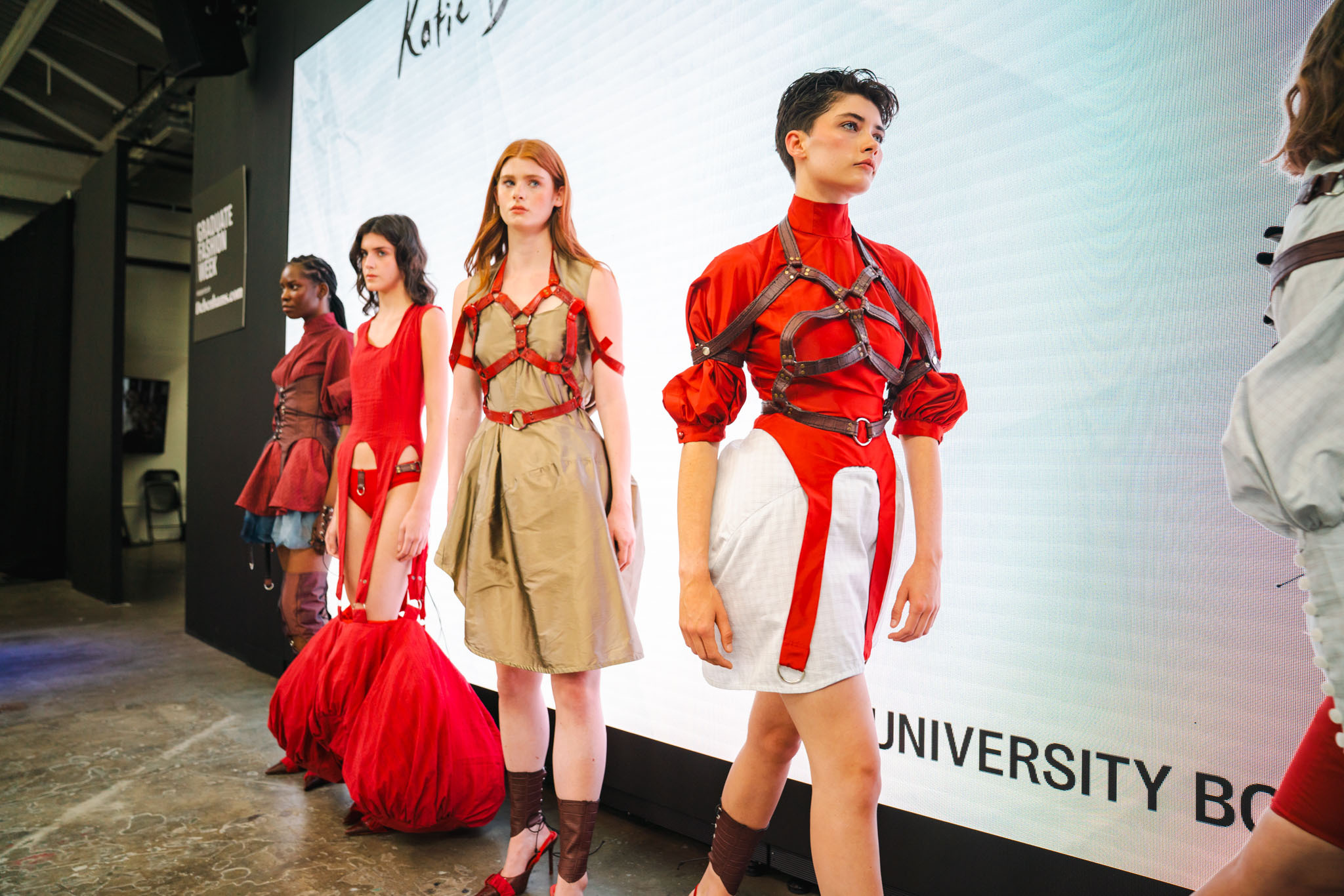 Fashion show models on runway wearing red and beige garments, with large screen backdrop displaying university branding.