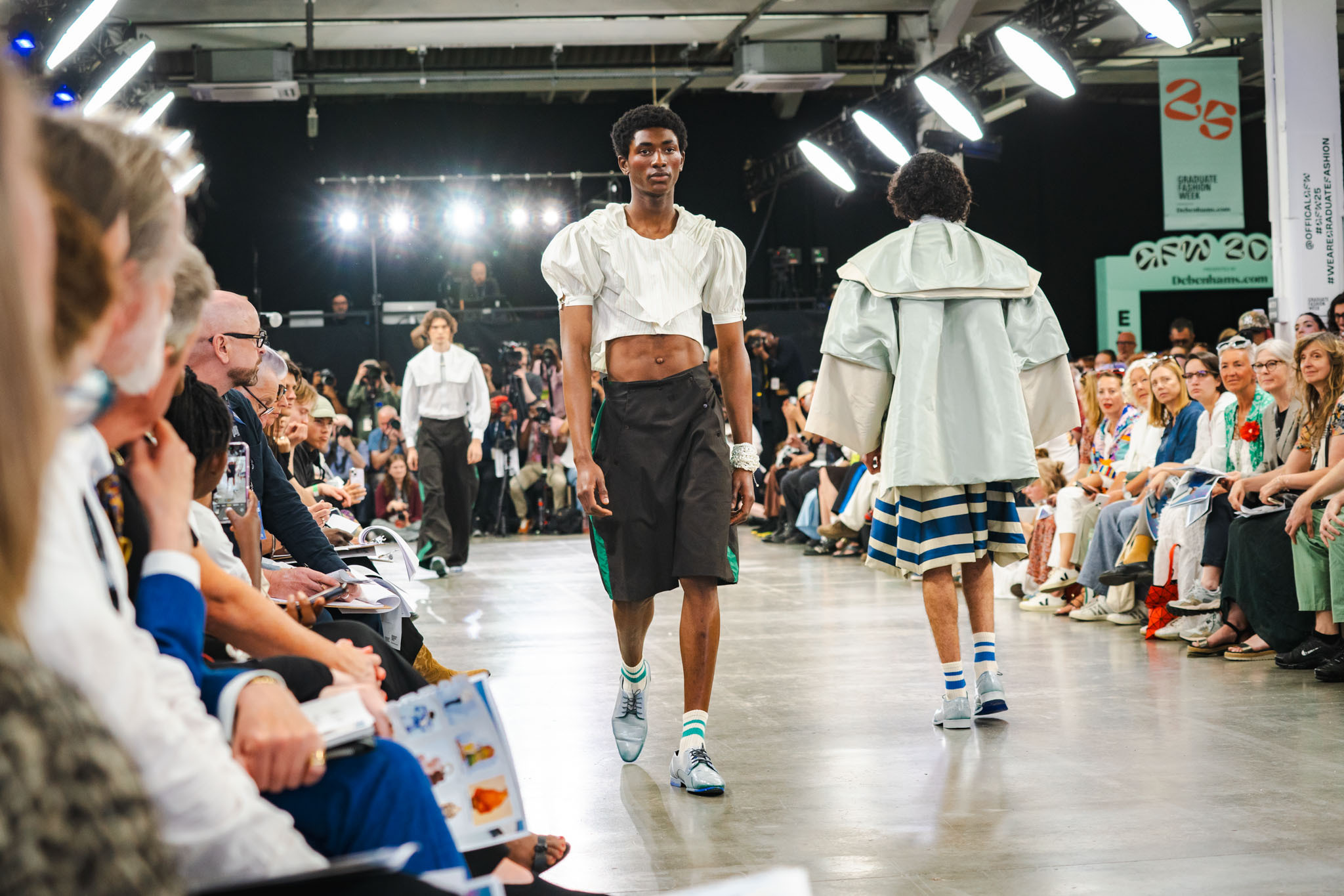 Models walking down runway in industrial venue with seated audience on both sides, wearing white tops and dark bottoms.
