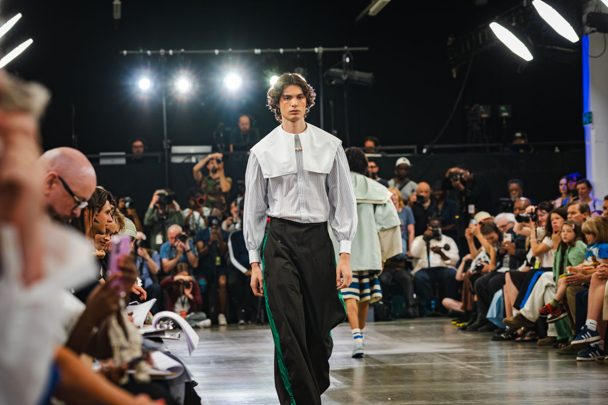 Male model walks down runway in white shirt and black trousers, surrounded by seated audience under bright stage lighting.