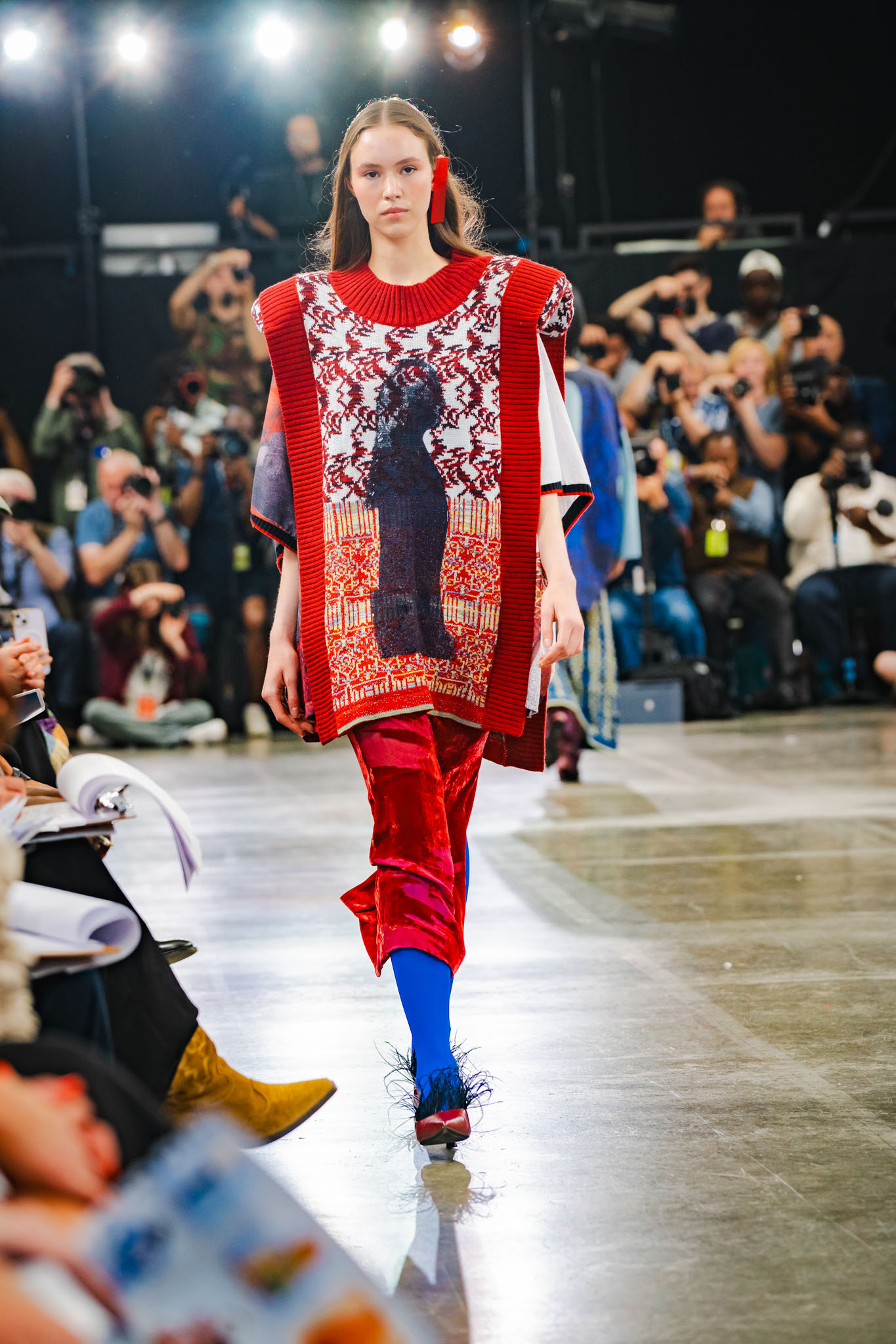 Model walks fashion runway wearing red oversized jumper with black cat motif, red trousers, blue tights. Audience and photographers visible.