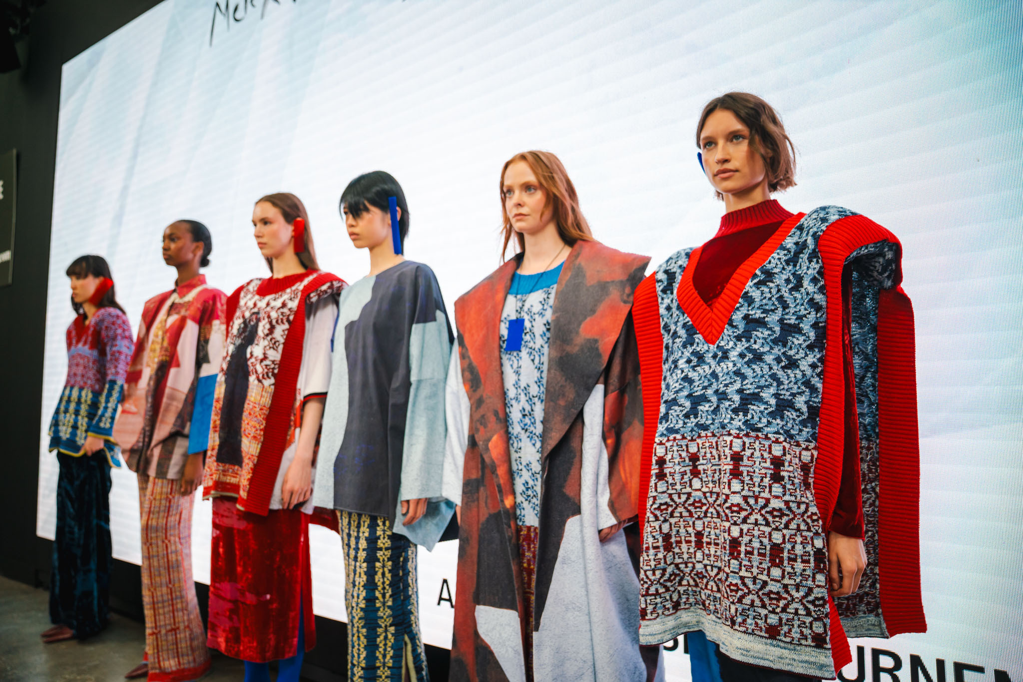 Seven models on catwalk wearing colourful knitwear and patterned garments in reds, blues, greys and whites at fashion show.