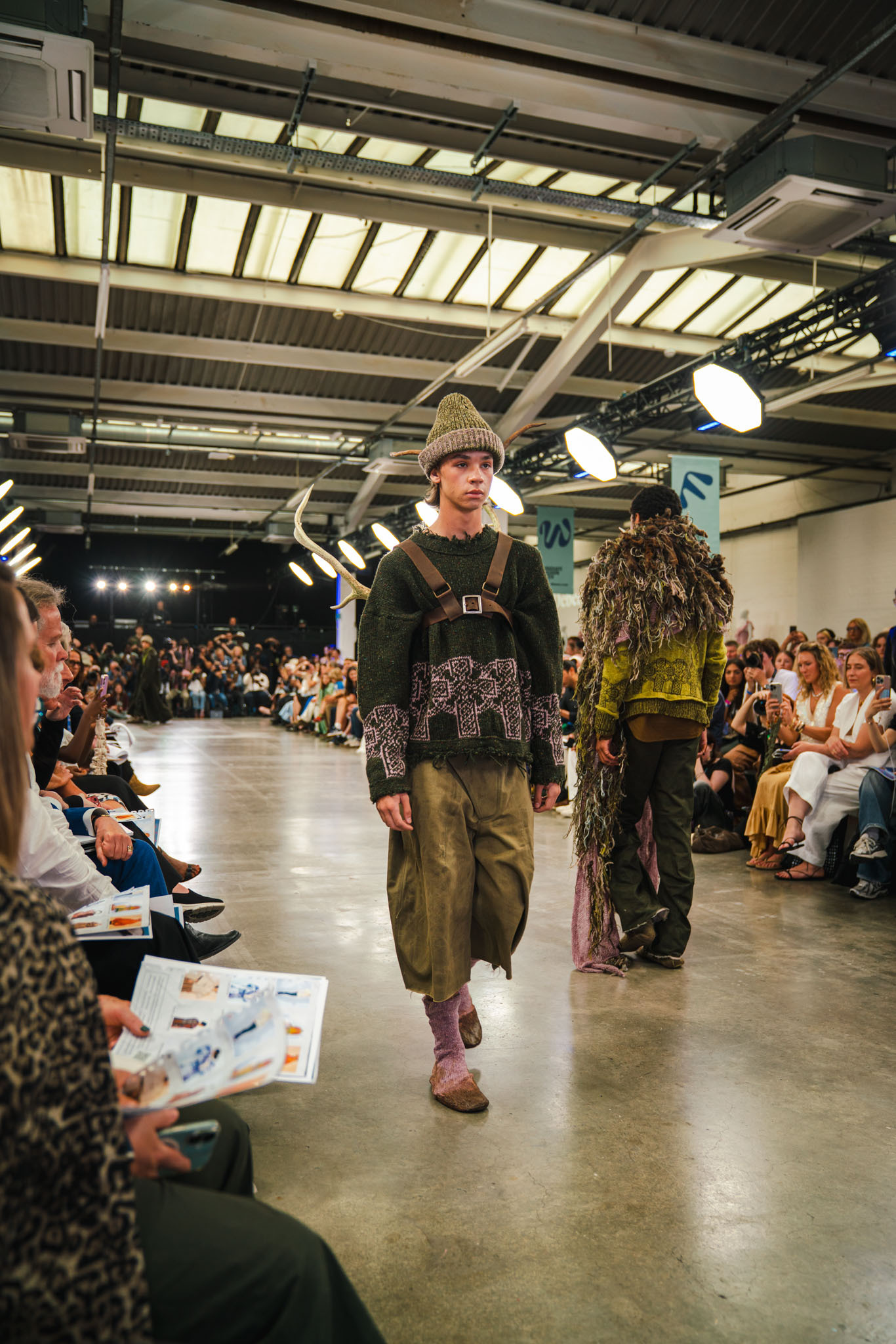 Male model in knitted hat and layered green outfit walks down runway in industrial venue with seated audience and exposed ceiling.