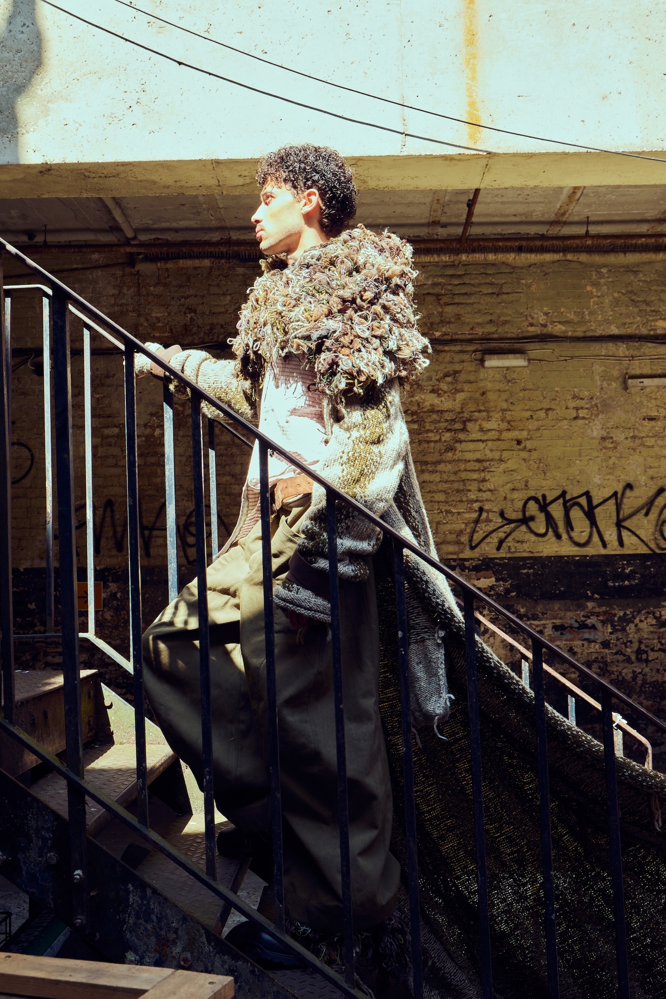 A model wearing a long, Medieval-style cloak with a fur collar walking up some stairs with metal railings running along the side.