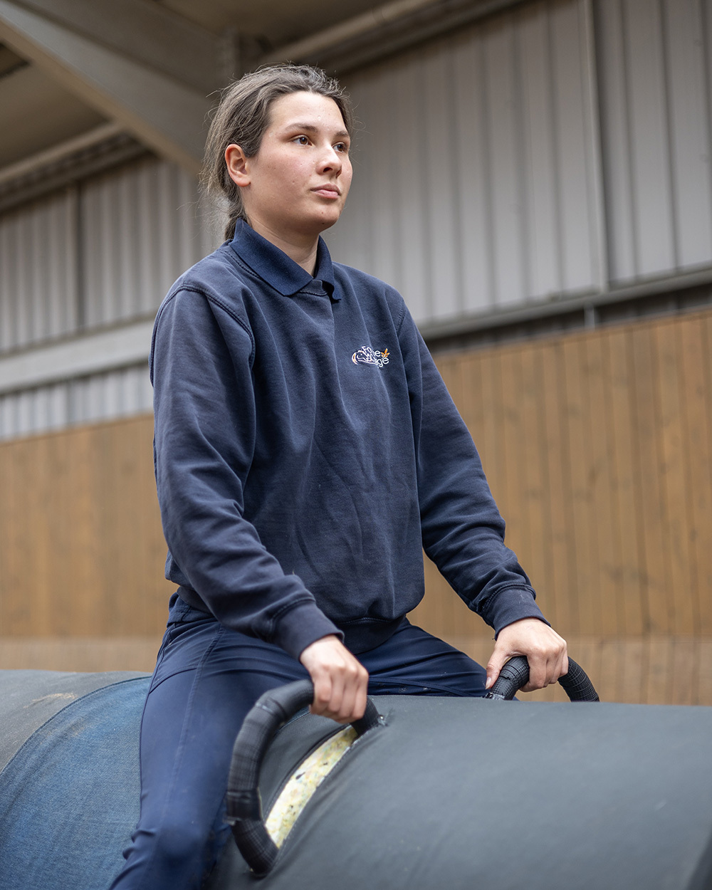 Woman in navy blue uniform sitting on a large cylindrical object in industrial warehouse setting with concrete pillars and wooden panels.