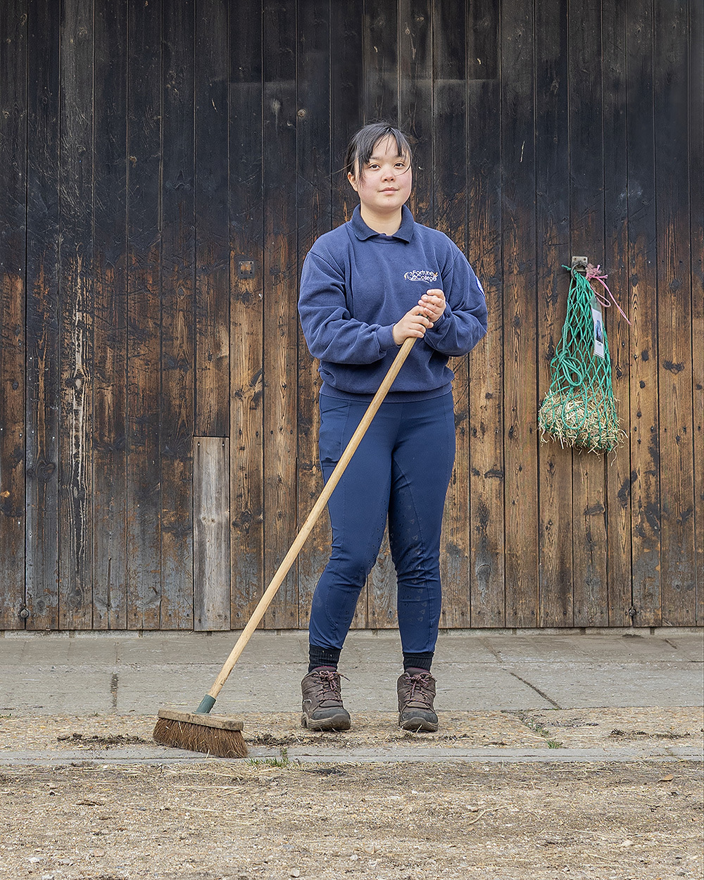 Woman in blue uniform holding wooden broom, standing on concrete ground against weathered wooden wall with green fishing net hanging.