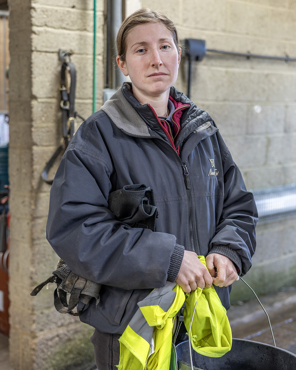 Woman in grey work jacket with hood up, holding bright yellow high-visibility clothing, standing in industrial setting.