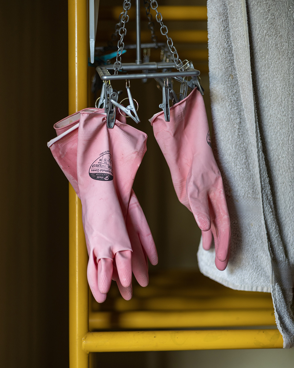 Pink rubber cleaning gloves hanging by clips on metal chain scale against yellow wooden surface and grey textured wall.
