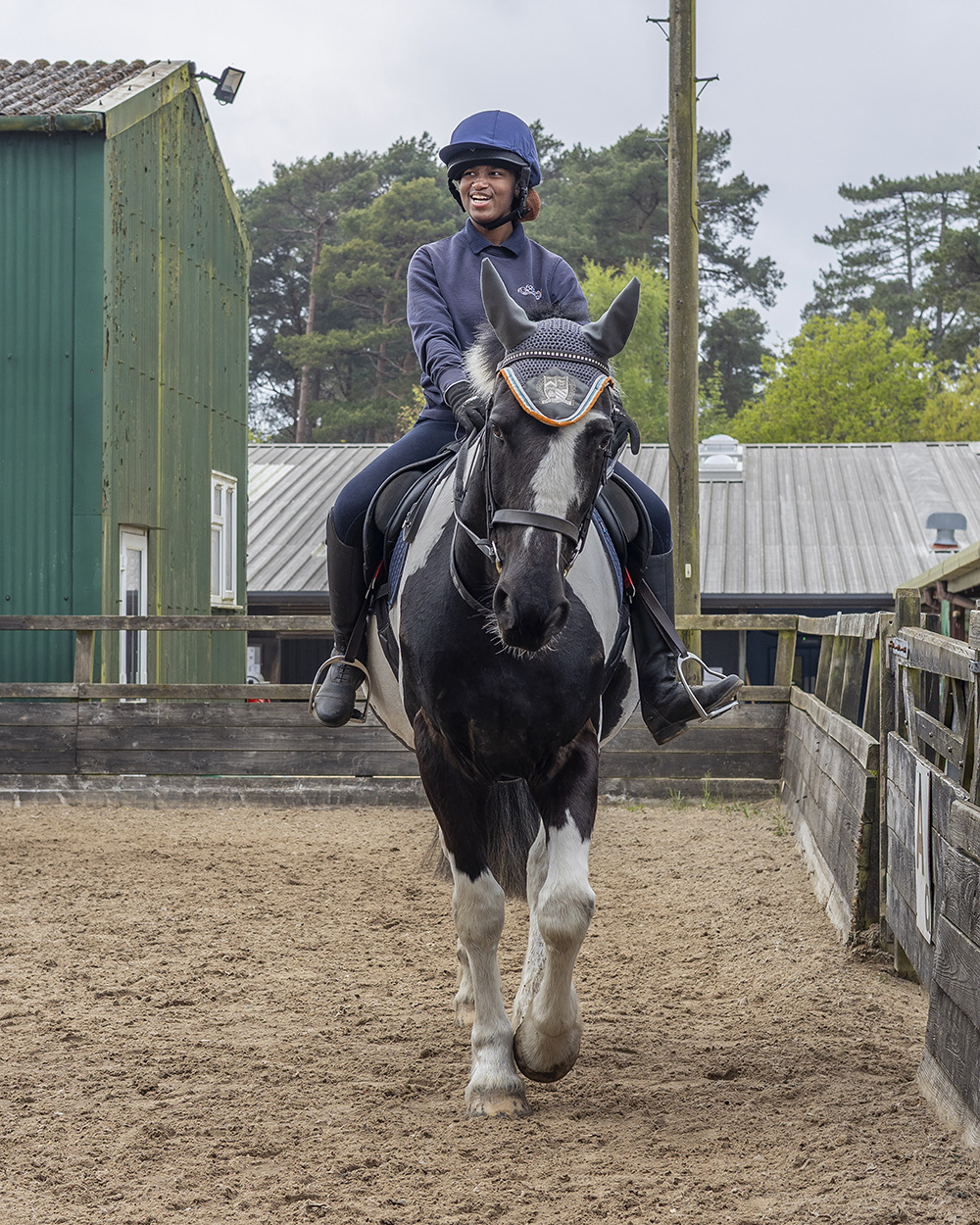 Rider in blue helmet and navy jacket on black and white horse trotting through stable yard with green buildings and wooden fencing.