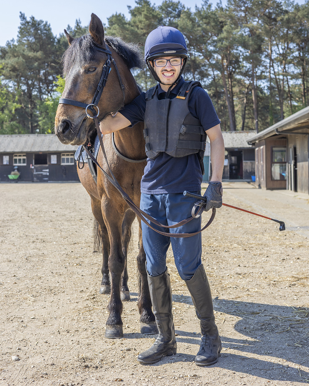 Young rider in navy helmet and protective vest standing beside brown horse with bridle in stable yard with trees behind.