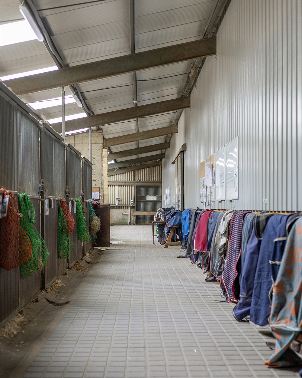 Industrial stable interior with metal stalls, concrete flooring, and colourful horse blankets hanging on rails under corrugated roof.
