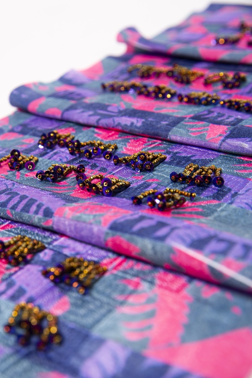 Purple and pink patterned fabric with gold sequined embellishments scattered across surface, fabric folded and draped on white background.