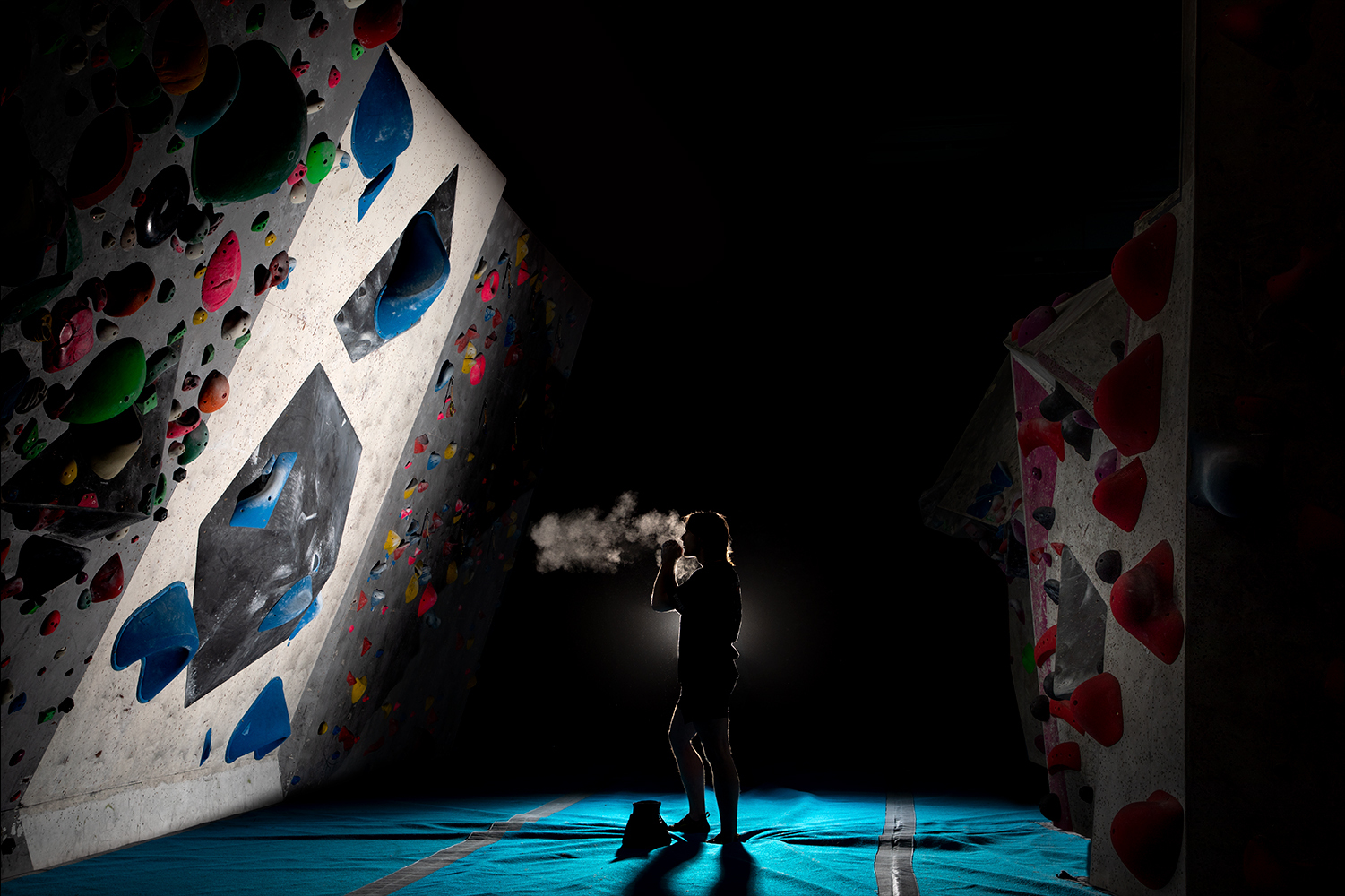 Person standing on blue-lit floor in dark room with colourful climbing wall featuring red, blue, green, and pink holds on white surface.