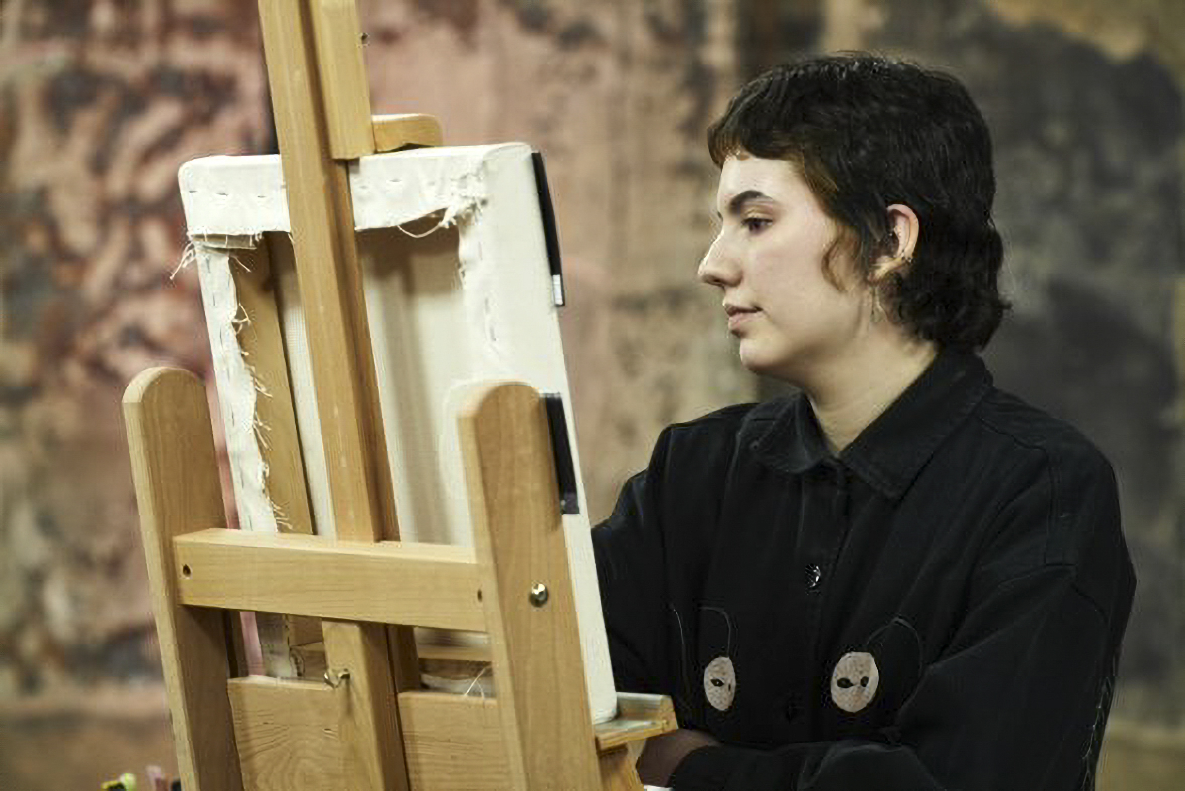 Artist G Cohen sits and works on a canvas on an easel, viewed from behind.
