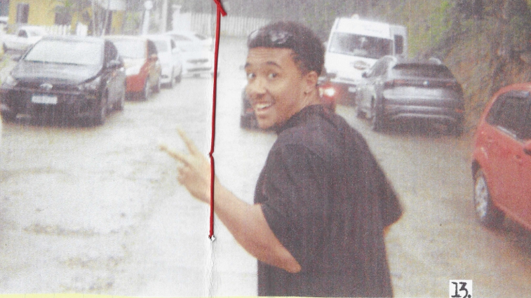 Scanned image of G Souza in Brazil, peace signing in the middle of a road by cars. The image has red thread through the middle and page numbers below.