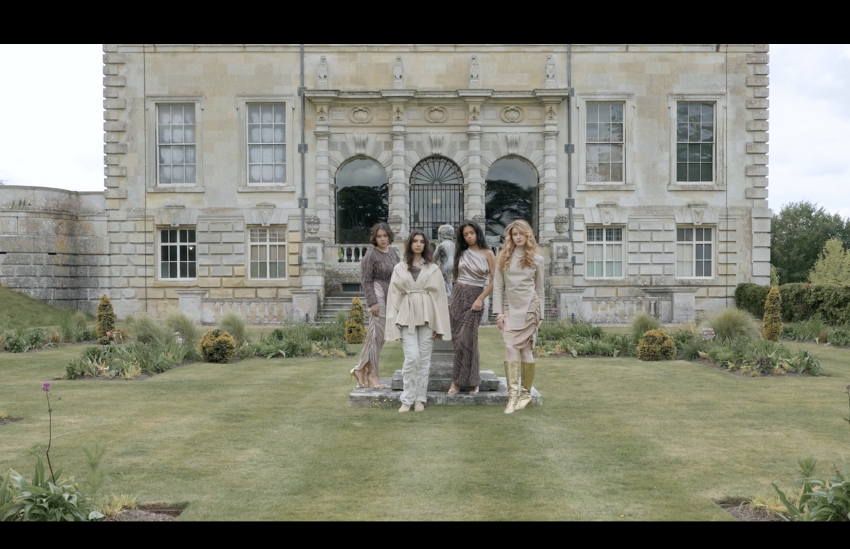Four women stand on a lawn in front of a grand stone mansion with arched windows and manicured garden borders.