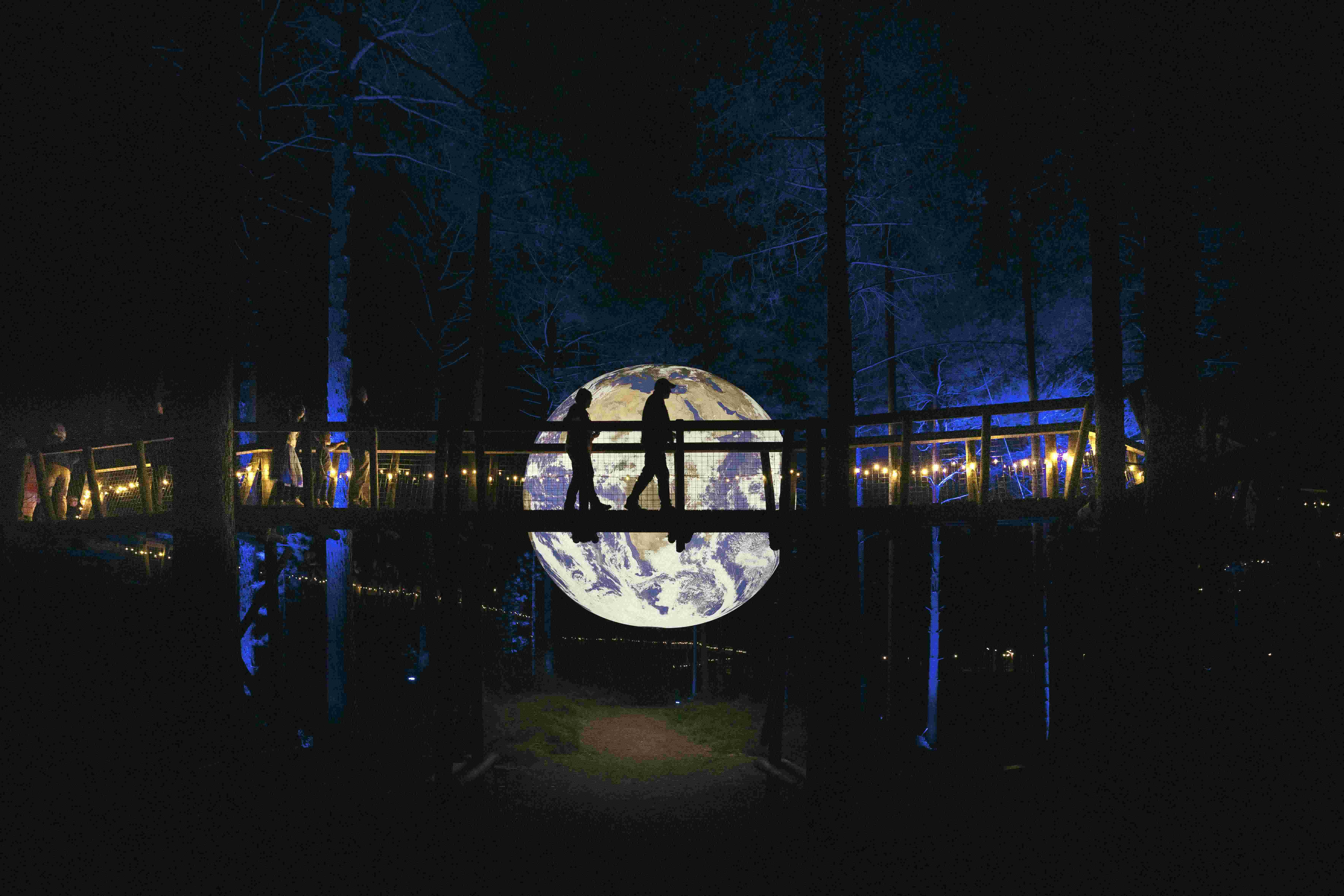 People cross a bridge set up around trees at night. A large glowing planet is visible in the centre.