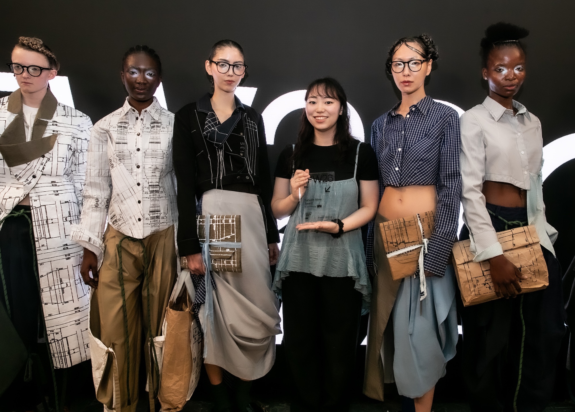 A group of six models stands together, showcasing various outfits designed with unique patterns and textures. The woman in the centre wears a black shirt and blue apron, while the others display an array of styles, including trousers and skirts paired with jackets.