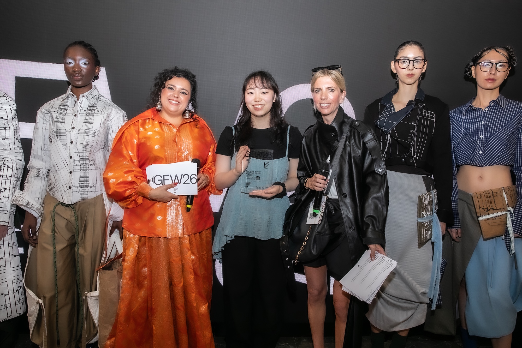 Five women stand together for a photograph, showcasing diverse fashion styles. One woman in an orange outfit holds a sign that reads 'GEW26'. The background is dark, accentuating their outfits.