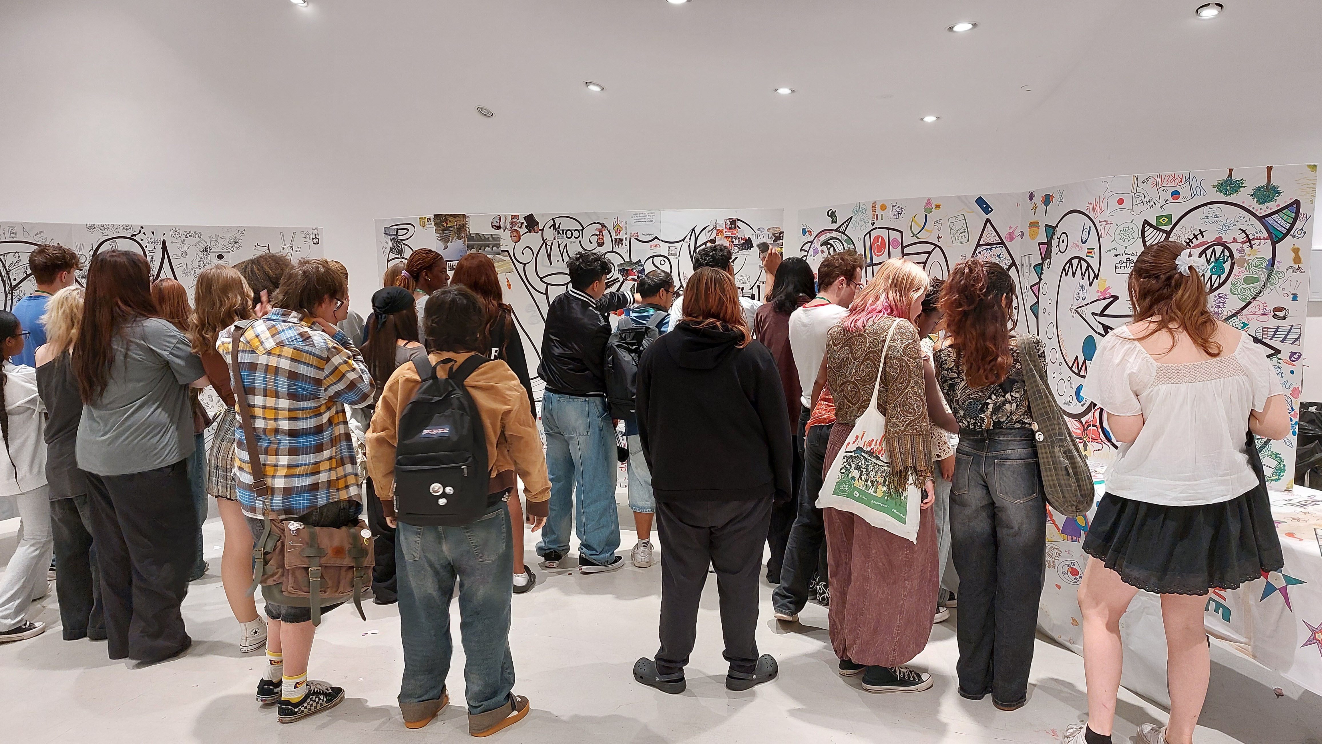 Crowd of people viewing art exhibition in white gallery space with drawings on walls and spotlights overhead.