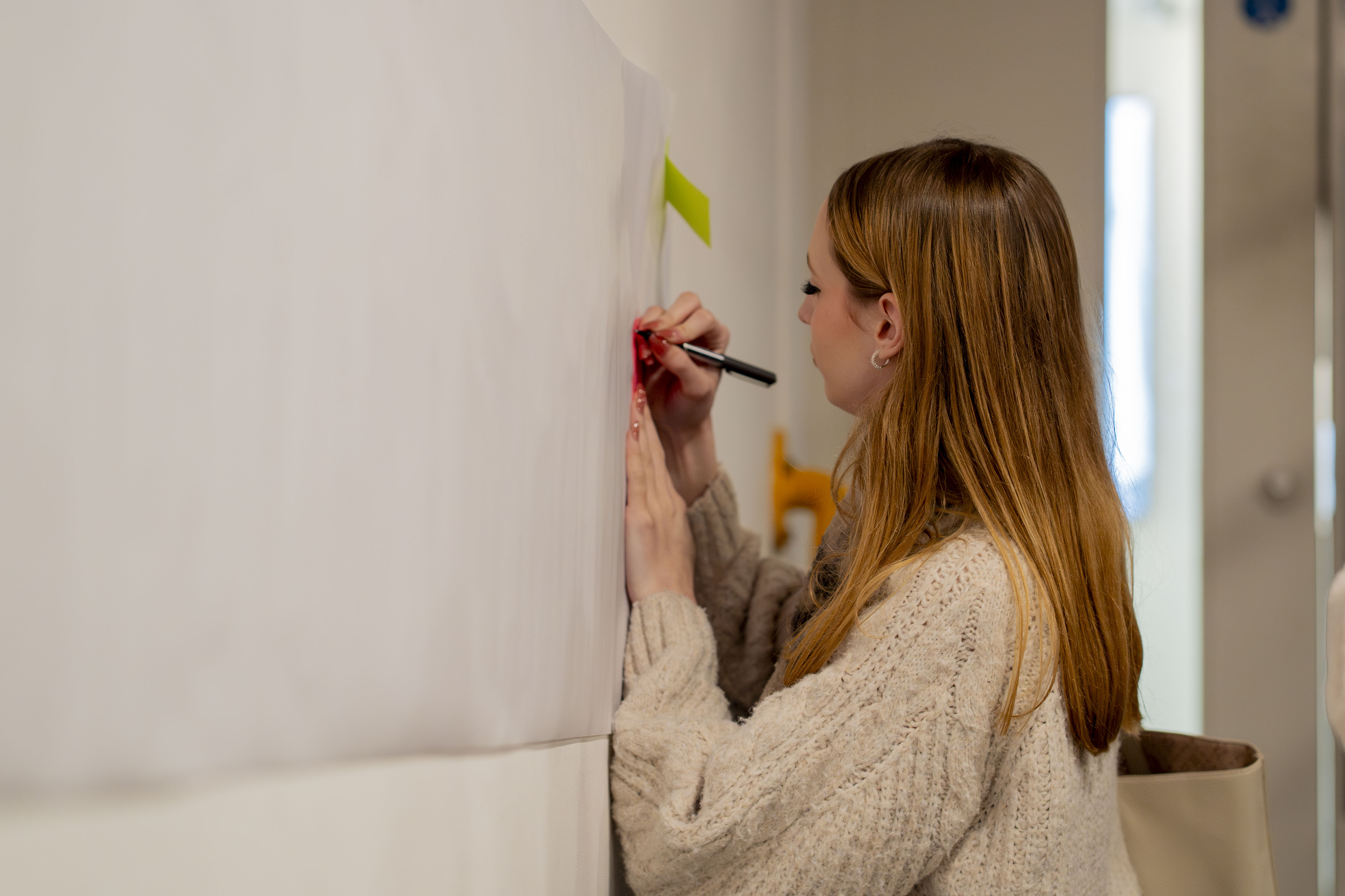 Woman with long blonde hair in cream jumper writing on white wall with marker pen in bright indoor space.