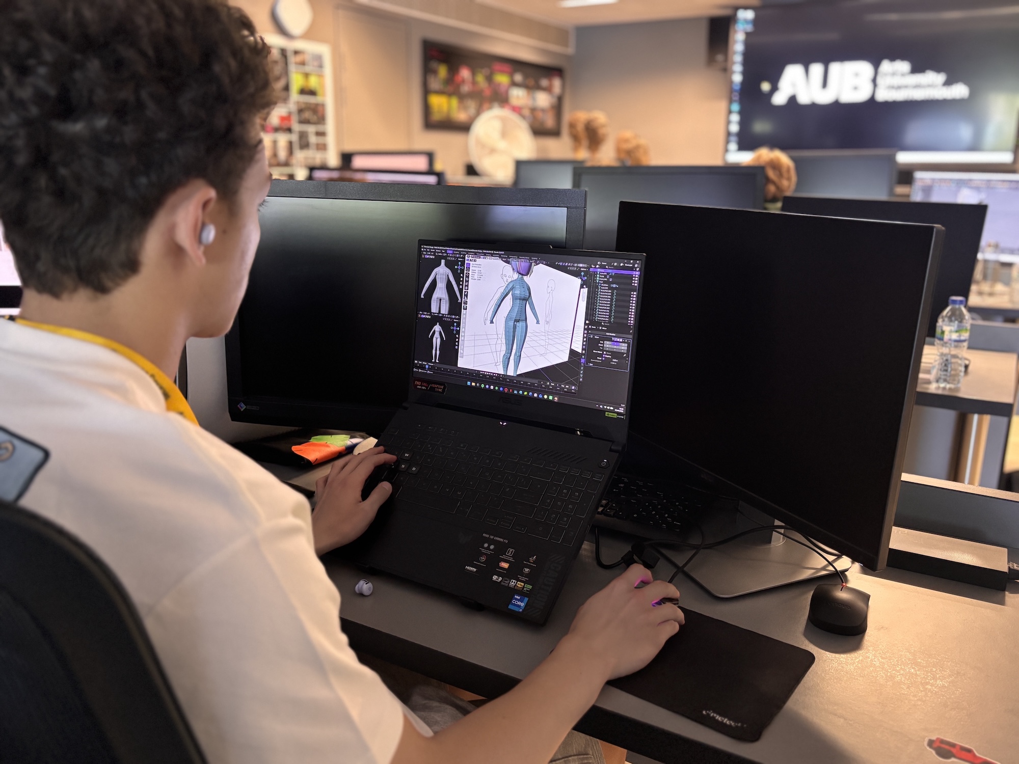 A person works at a laptop displaying 3D character design software, with dual monitors on a desk in a modern office or studio.
