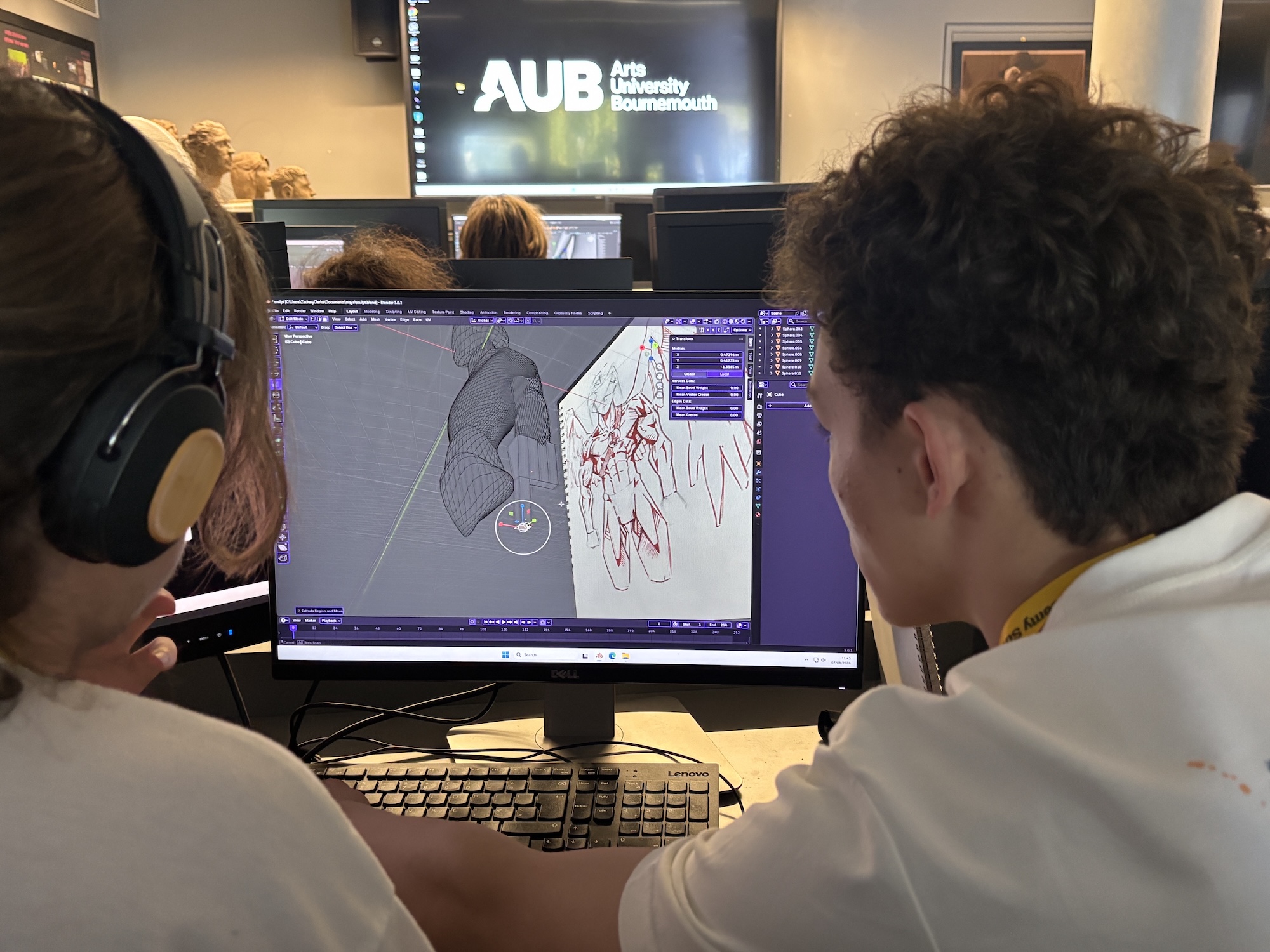 Two students wearing headphones work at a desktop computer displaying a 3D modelling interface, in a computer lab with an AUB Arts University Bournemouth screen in the background.